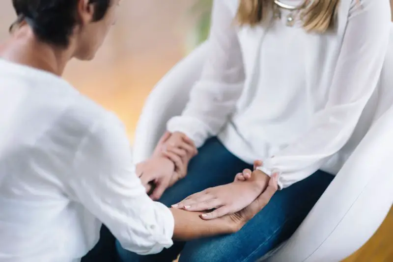 Female Theta healing therapist performing alternative therapy treatment with young woman patient. Therapist holding hands and transfer energy. Wearing white clothes.